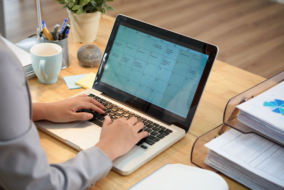 Close-up of hands on a laptop keyboard on a wooden table, with a monthly calendar and highlighted date ranges on the screen.