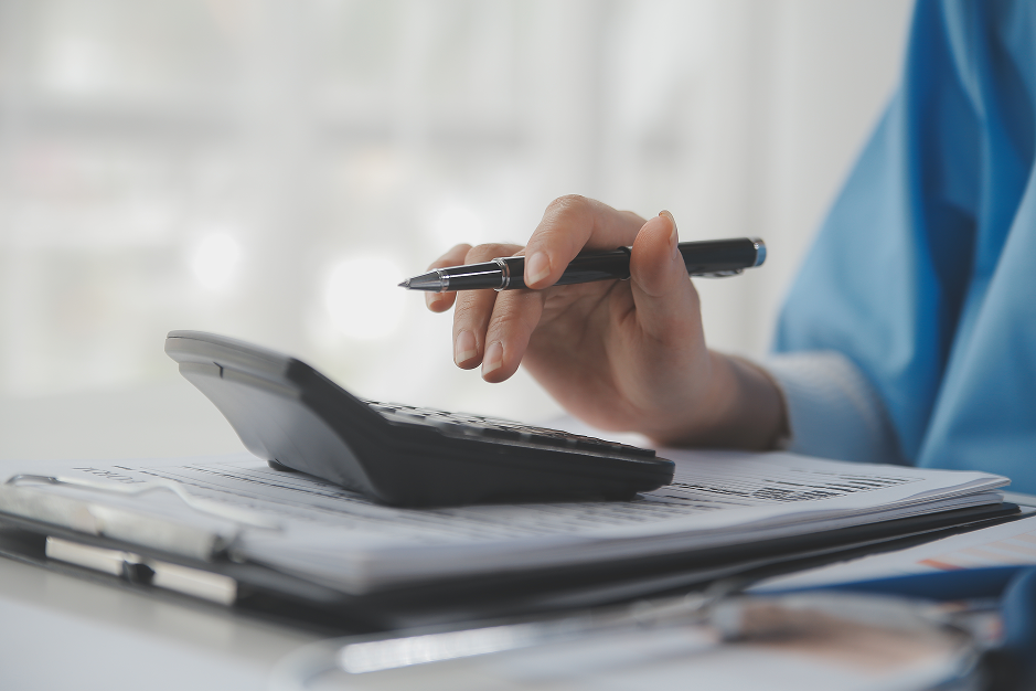 A person in blue clothing holds a black pen above a gray calculator resting on stacked documents and folders. The photograph focuses on the hand, calculator, and paperwork; no title is overlaid.