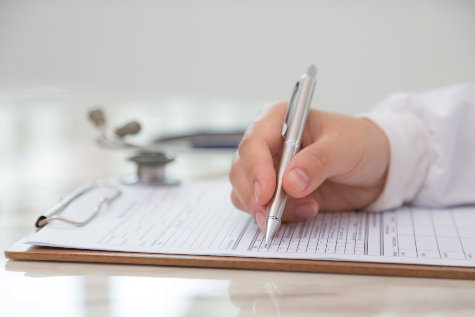 A person in a white coat writes on a medical form clipped to a board, with glasses and a stethoscope on the desk.