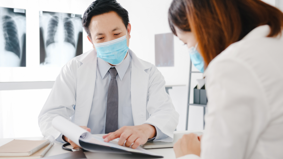 A male doctor wearing a white coat and blue surgical mask points to documents on a desk while a woman seated across from him looks at the paperwork. X-ray images are visible behind the doctor.