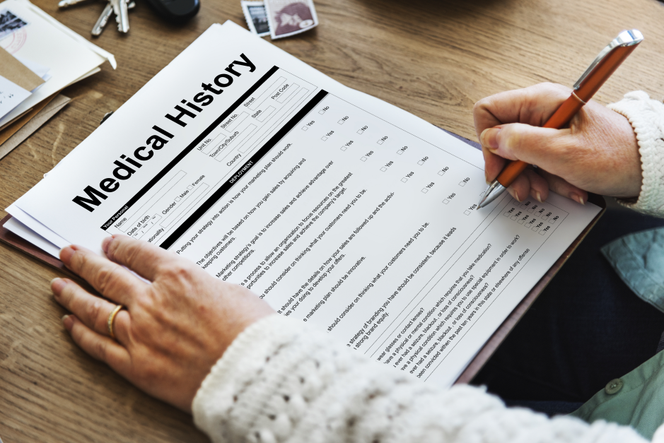 An overhead photograph of a person holding a medical history form on a clipboard and writing on it with a pen at a wooden desk.