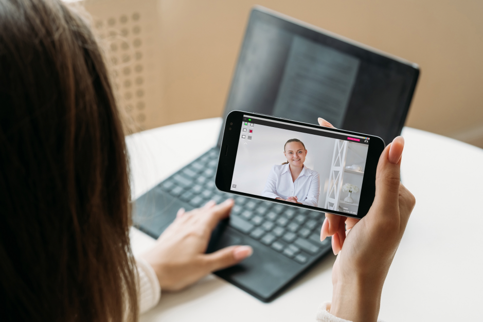 Viewed from behind, a woman holds a smartphone horizontally during a video call with another woman wearing a white shirt. An open laptop sits on the table behind the phone.