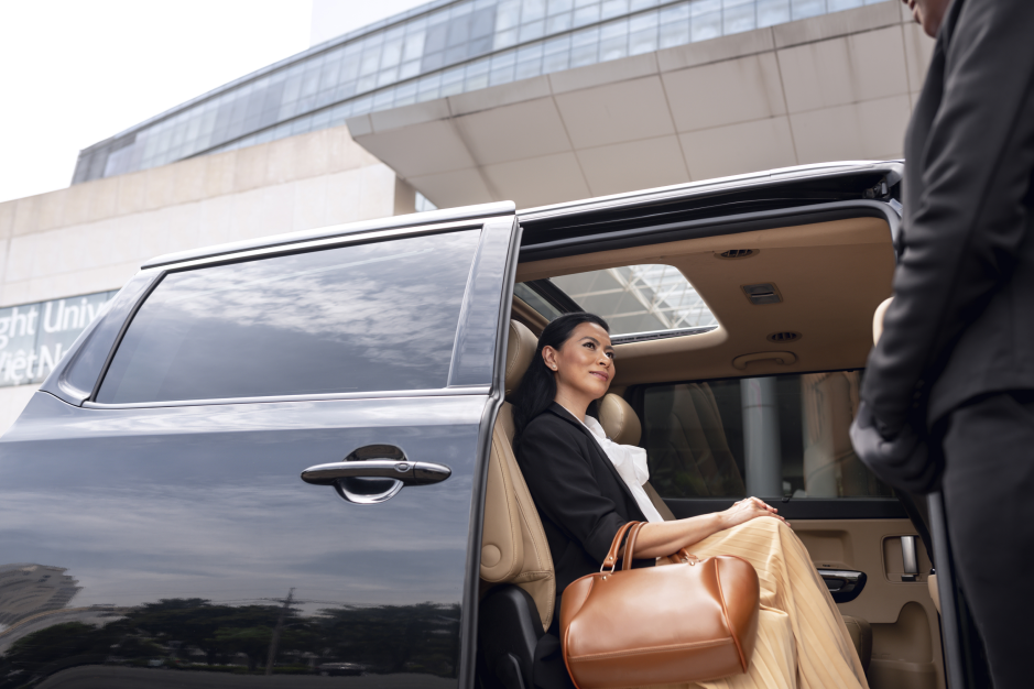 A woman with a brown handbag sits in the rear seat of a dark vehicle with its door open. Another person stands beside the vehicle, with a large building in the background.