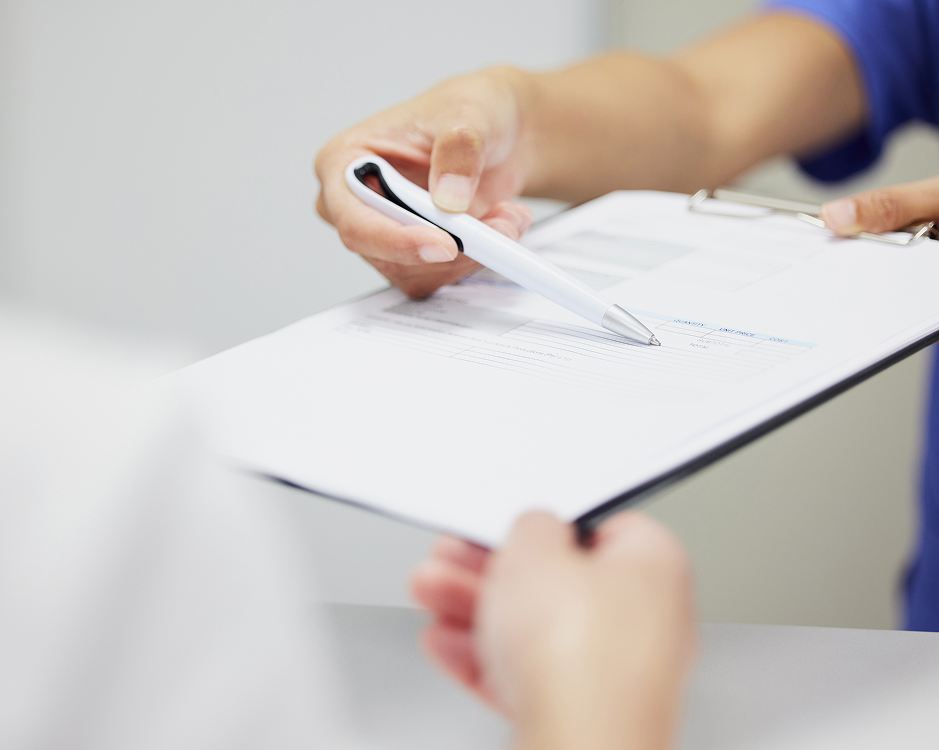 Close-up of two people's hands around a black clipboard with white paperwork. One hand holds a silver pen pointing toward the document, while another grips the clipboard. Blue clothing is visible in the background; no title is overlaid.