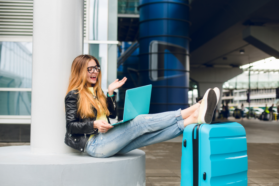 A woman wearing glasses and a black jacket sits by an airport column with a turquoise laptop on her lap and her feet resting on a matching suitcase.