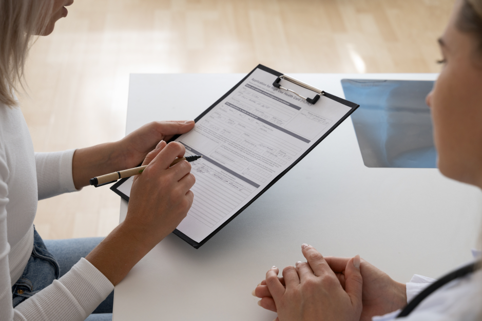 An overhead view of a clinician holding a clipboard and pointing a pen at a printed form while a seated patient rests her clasped hands on the table.