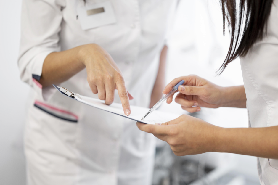 Two people in white uniforms review a medical form on a clipboard, with one pointing to an entry and the other holding a pen.