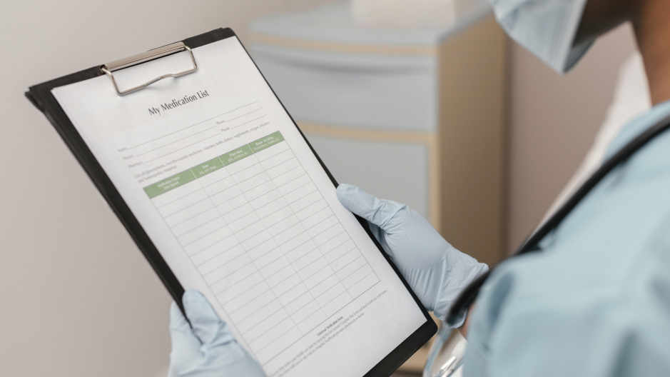 A healthcare worker wearing gloves holds a clipboard labeled “My Medication List” and reviews its entries.
