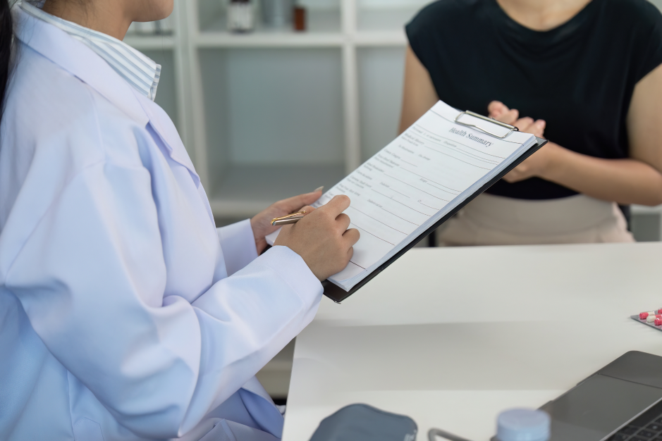 A cropped photograph of a doctor in a white coat holding a clipboard and a pen across a desk from a seated patient wearing a black top.