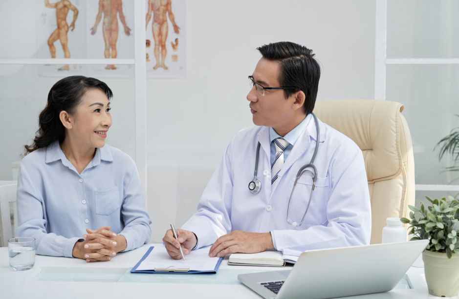 Doctor holding a pen over a clipboard while speaking with a woman across a clinic desk, illustrating the article’s review of consultation plans and appointments before travel to Korea