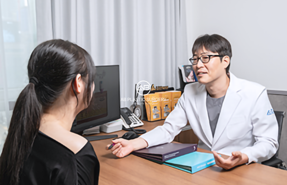 A clinician wearing glasses and a white coat speaks with a seated patient across a desk; a monitor, telephone and folders are visible, with no price displayed.
