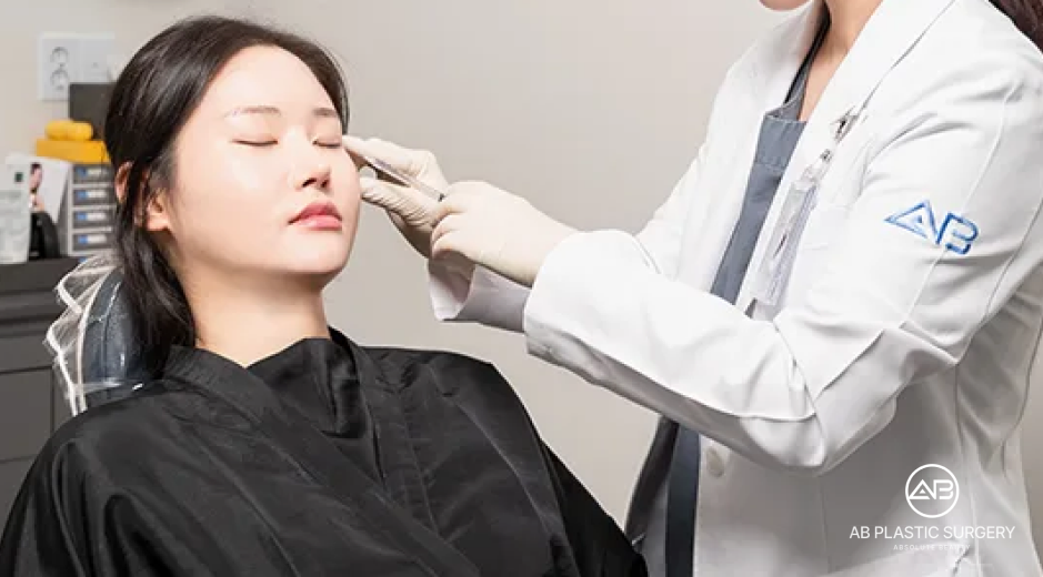 Seorang perempuan duduk dengan mata tertutup saat dokter berjas putih memeriksa area sekitar mata dan pipinya menggunakan tangan bersarung. Logo AB PLASTIC SURGERY terlihat di kanan bawah.