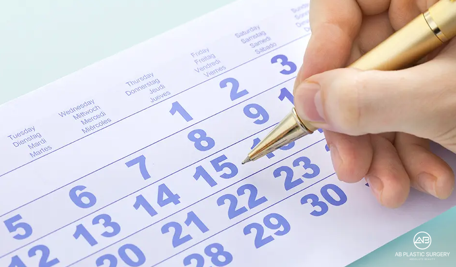 Foto dekat tangan memegang pena di atas kalender kertas dengan tanggal berwarna biru. Logo AB terlihat di pojok kanan bawah; gambar tidak menampilkan pasien atau hasil operasi.