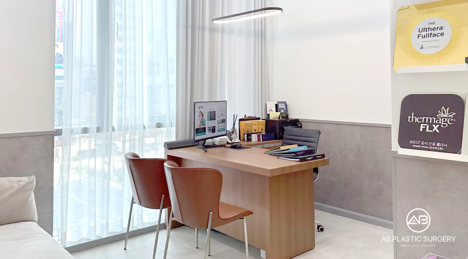 Ruang konsultasi dengan meja kayu, kursi pasien, monitor, lampu pemeriksaan, dan jendela bertirai tipis. Poster Ultherapy dan Thermage FLX terlihat di dinding kanan; tidak ada pasien dalam foto.